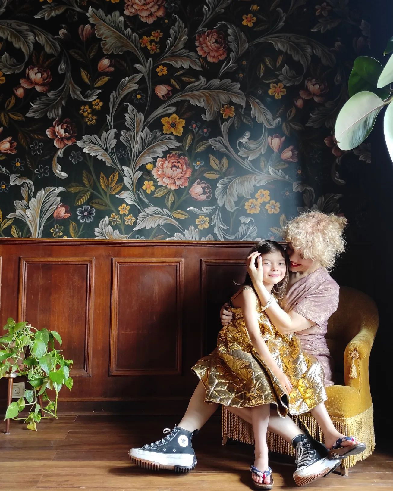 A woman and child pose in a chair against floral wallpaper. The child wears a gold dress, and the woman wears a pink dress and black Converse sneakers.