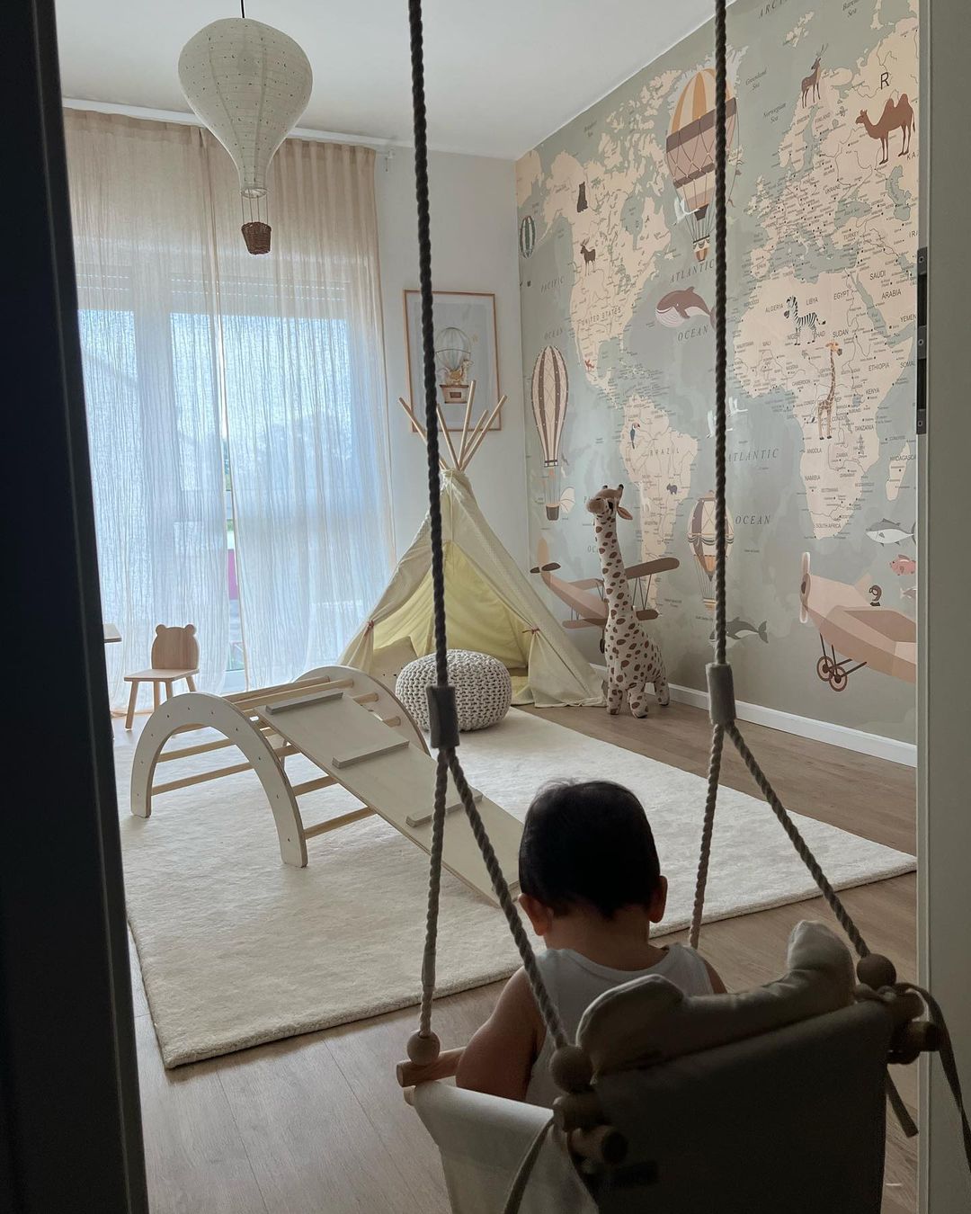 Bambino sull'altalena in una stanza dei giochi dai toni neutri con carta da parati con mappa del mondo, tenda e struttura per arrampicarsi.