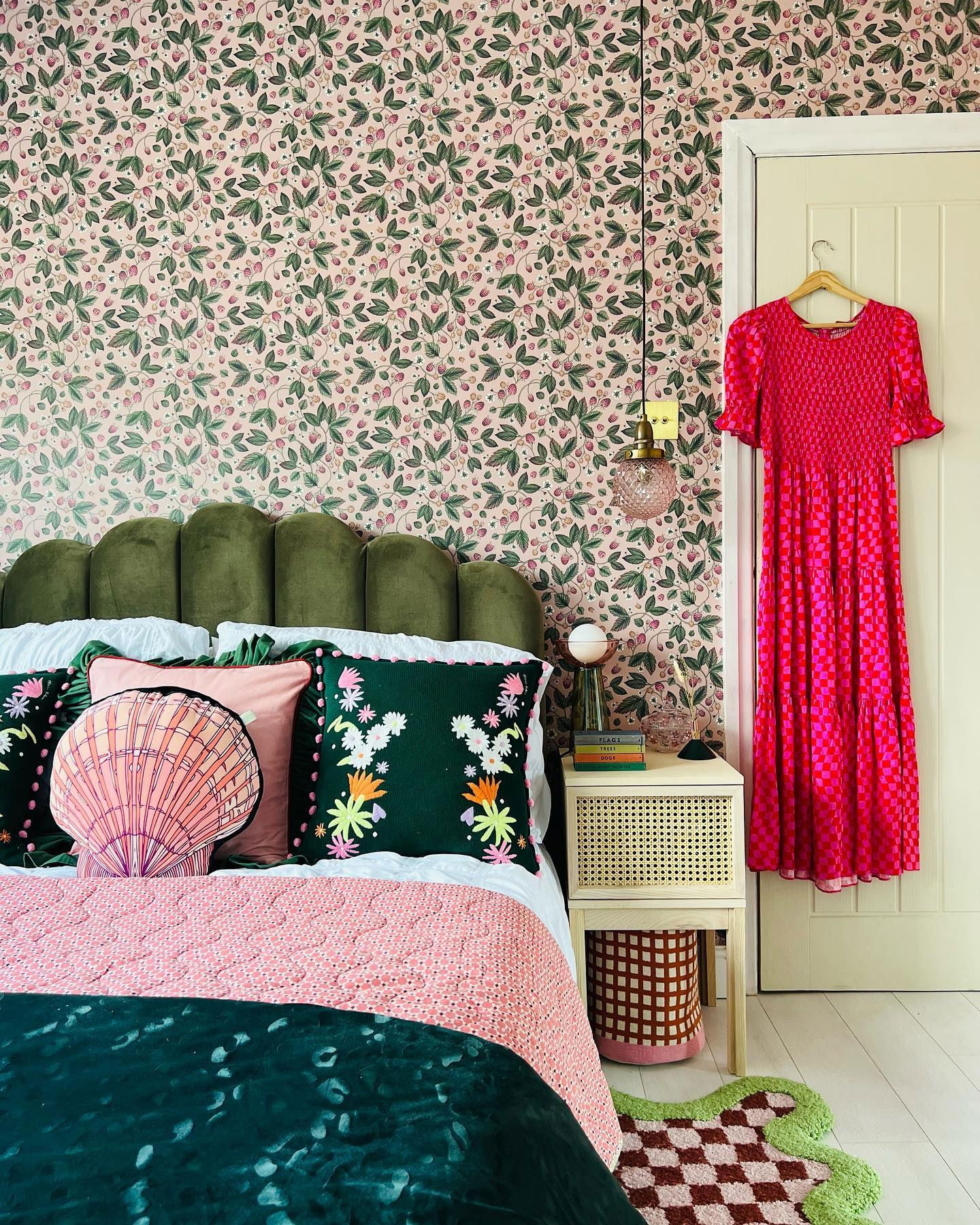Eclectic bedroom with pink floral wallpaper, green velvet headboard, and colorful pillows. A pink dress hangs on the door.
