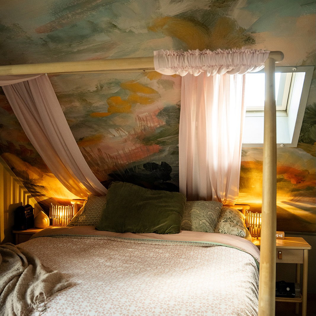 Ein gemütliches Schlafzimmer mit einem Himmelbett, das mit durchscheinendem rosa Stoff drapiert ist. Ein lebendiges abstraktes Wandgemälde bedeckt die abgeschrägte Decke und die Wände, beleuchtet von warmen Nachttischlampen.