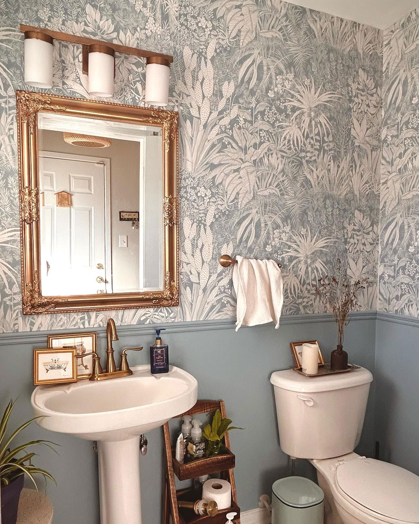 Bathroom with blue botanical wallpaper, gold mirror, pedestal sink, and toilet with decor.