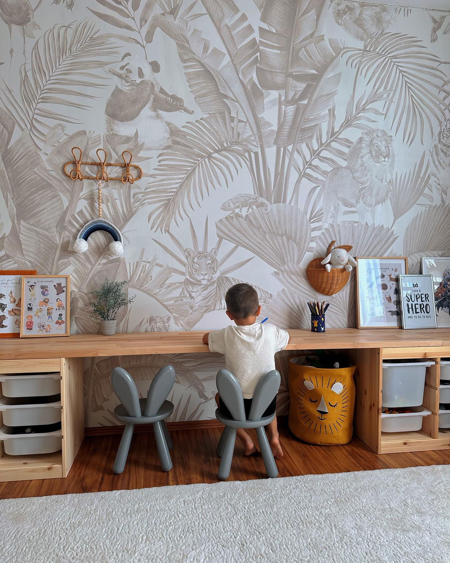 Child sits at wooden desk with gray bunny chairs. Jungle-themed wallpaper, lion storage bin, and white rug.