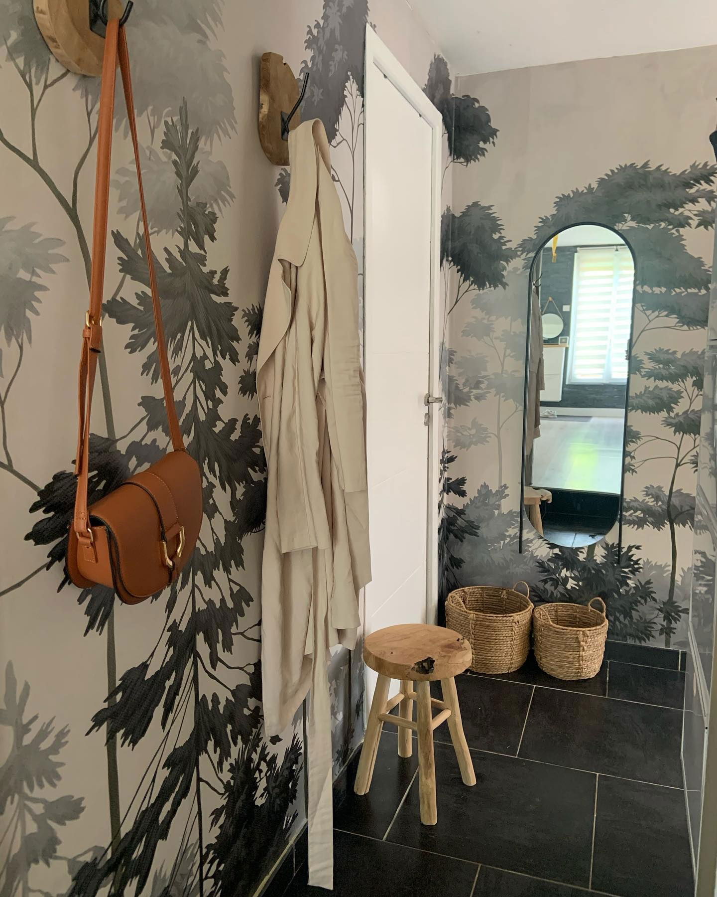 Hallway with forest wallpaper, black tile floor, and decor. A brown purse and beige coat hang on wooden wall hooks.