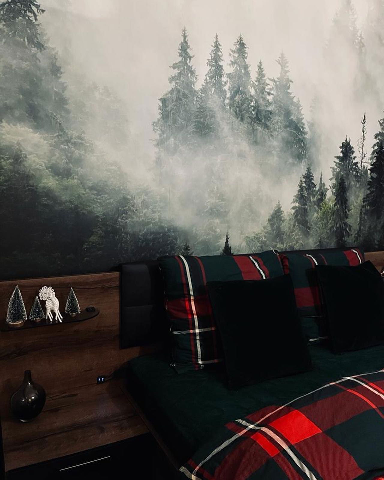 Schlafzimmer mit Waldgemälde, karierter Bettwäsche und dekorativen Bäumen.