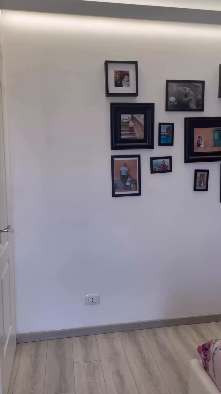 White wall with black-framed family photos, light wood floor.