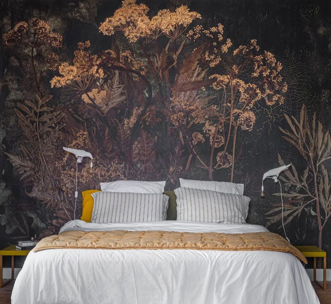 Schlafzimmer mit weißer Bettwäsche, gestreiften Kissen und einer goldenen Steppdecke. Blumentapeten und vögelförmige Wandleuchten setzen einzigartige Akzente.