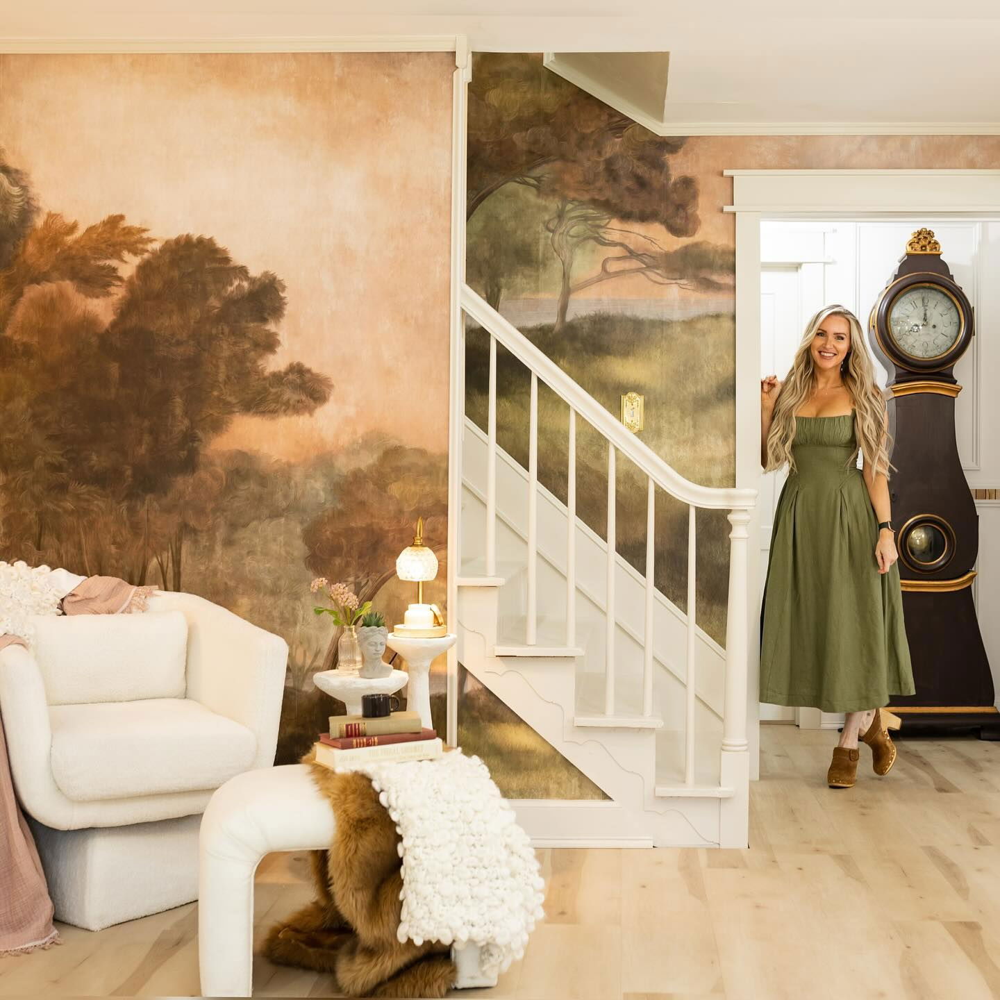 Living room with landscape mural, white chair, and woman in green dress standing near a tall clock.