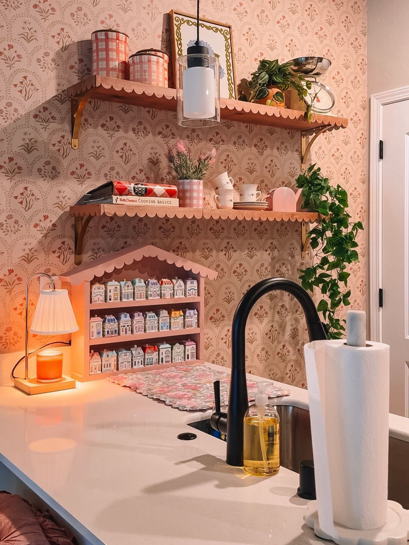 Estantes rosas festoneados con decoración y plantas contra un papel pintado floral. Un mostrador tiene un grifo negro, toallas de papel y una exhibición rosa en forma de casa llena de pequeños frascos con forma de casa junto a una lámpara de vela brillante.
