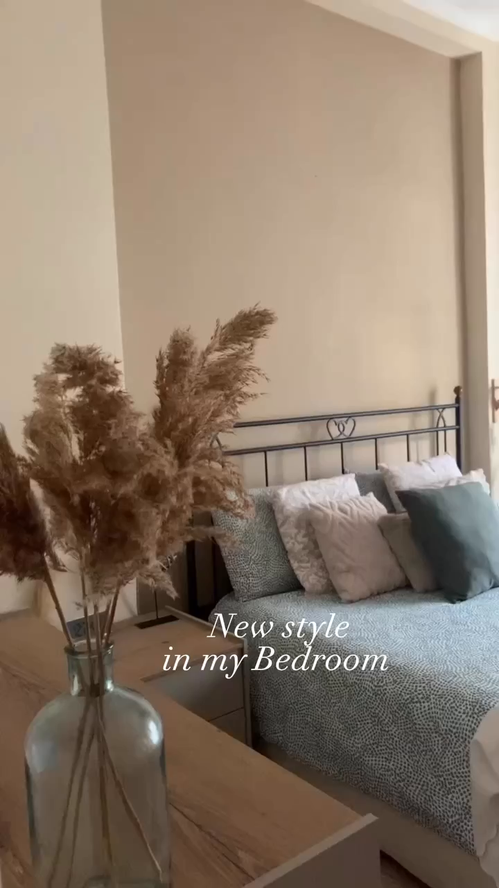 Bedroom interior with pampas grass in a glass vase on a wooden nightstand. Bed has a black metal frame and patterned bedding.