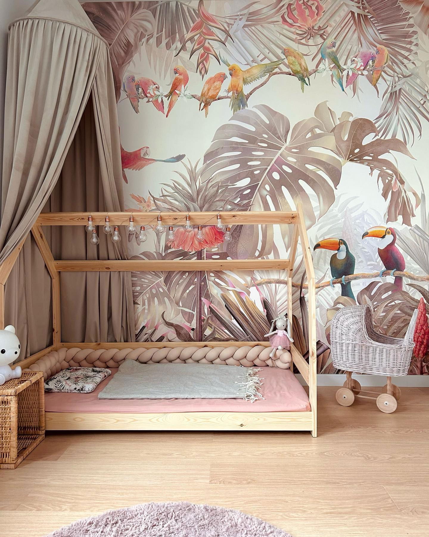 Child's bedroom with tropical wallpaper, wooden bed frame, braided bumper, and wicker doll stroller.