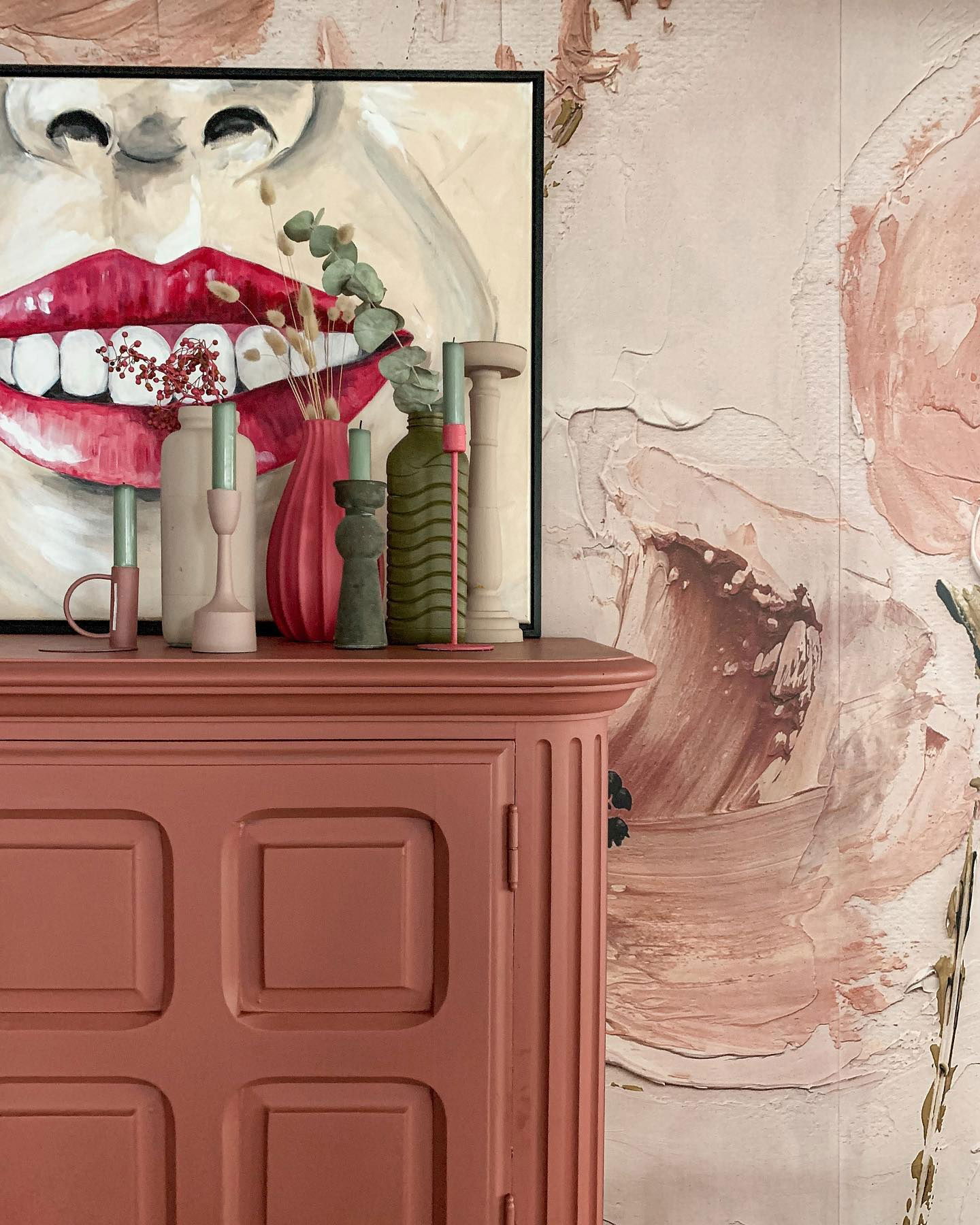 Close-up of pink cabinet with art and decor. Painting of mouth with red lips and dried floral arrangement. Pink floral wallpaper.