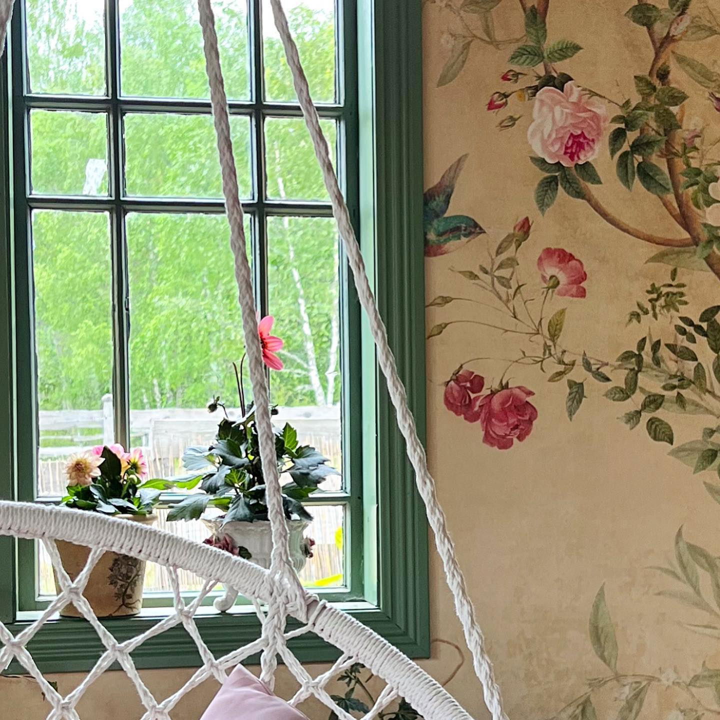 Interiørvisning af en hvid macrame-gyngestol nær et vindue med blomstret tapet.