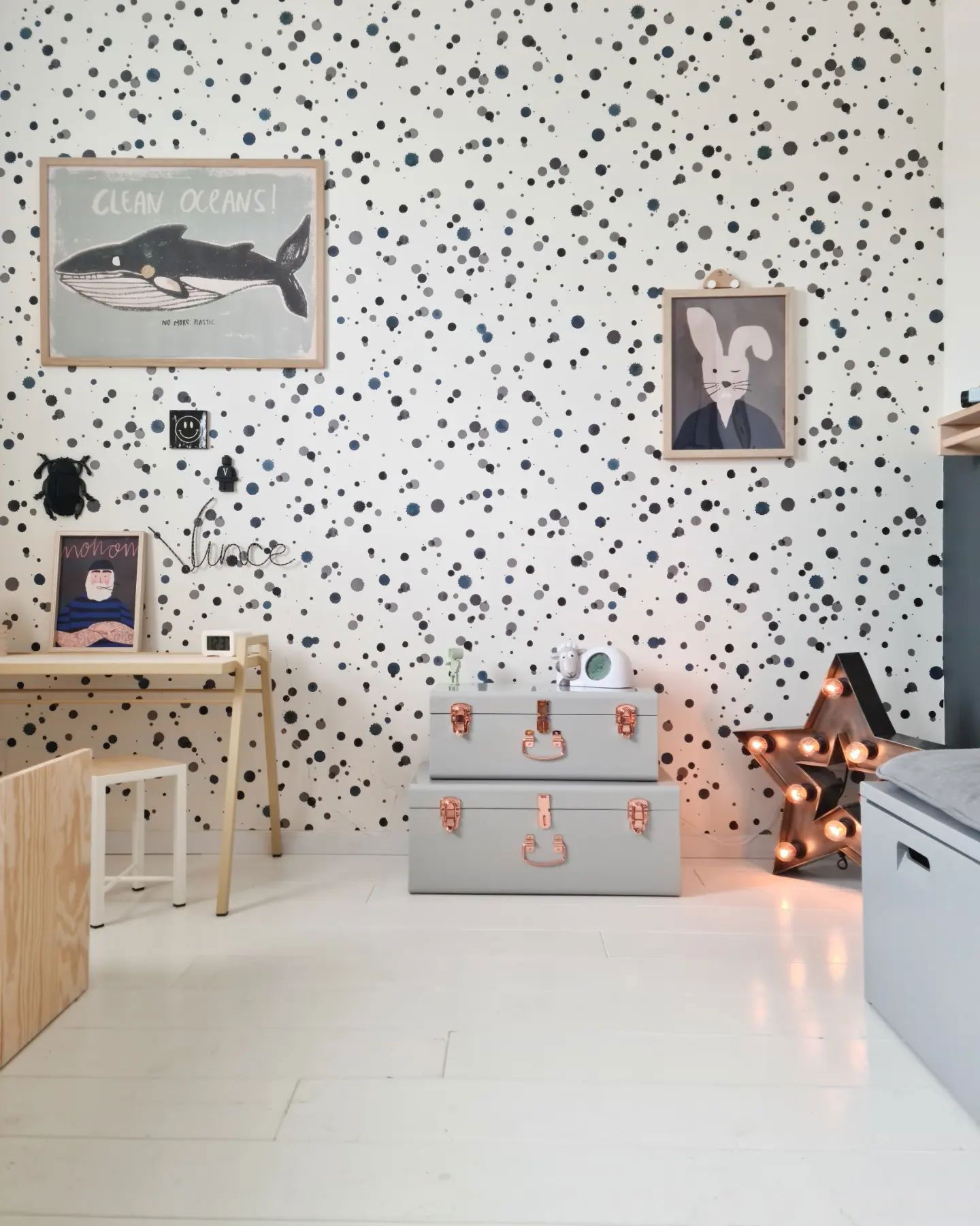Child's room with desk, polka dot wallpaper, and framed art. Two gray trunks with copper hardware are stacked near a lighted star.