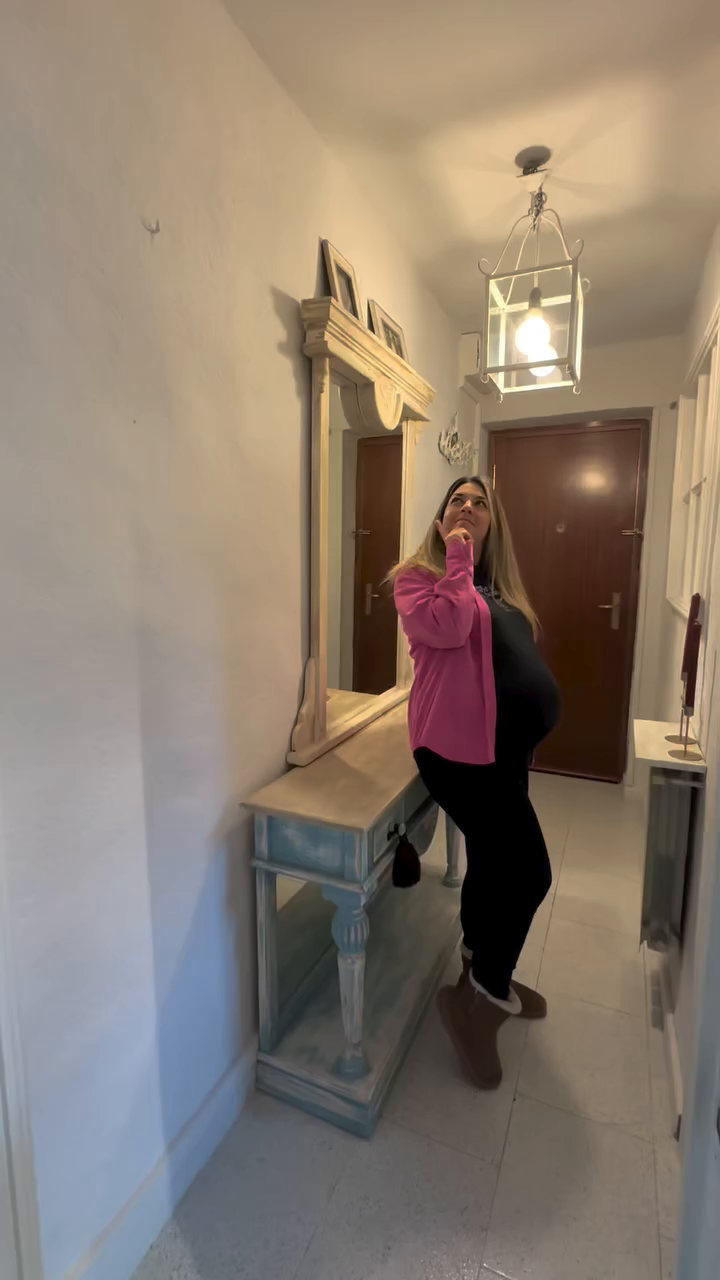 A pregnant woman in a pink cardigan stands in a hallway, looking up thoughtfully.