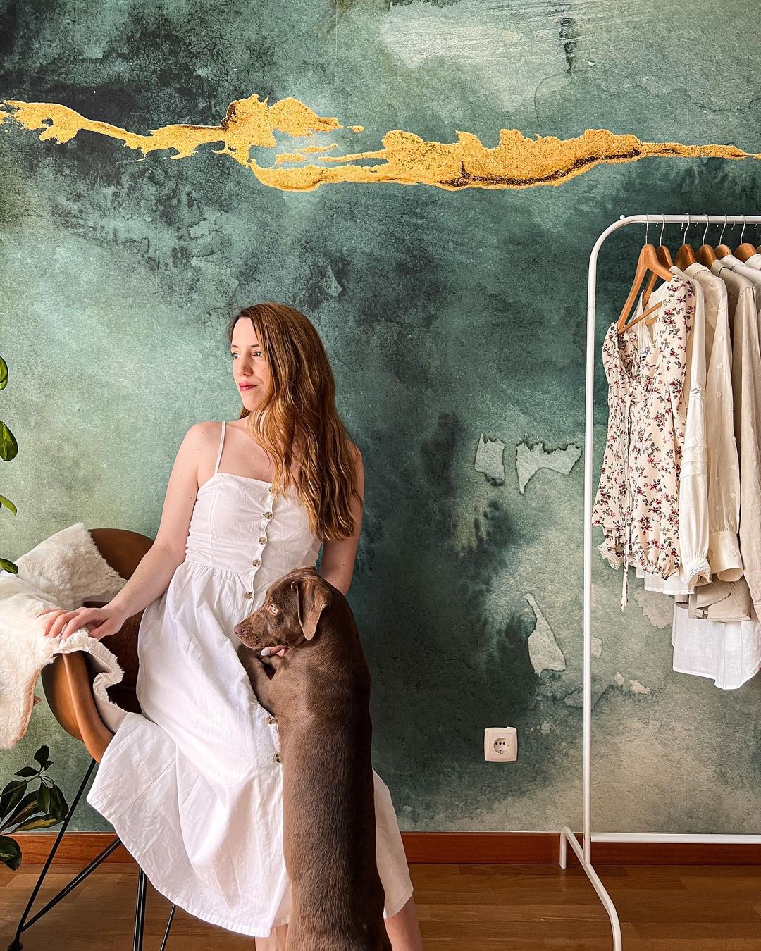 Een vrouw in een witte jurk zit met haar hond voor een kledingrek en een groen-gouden accentmuur.