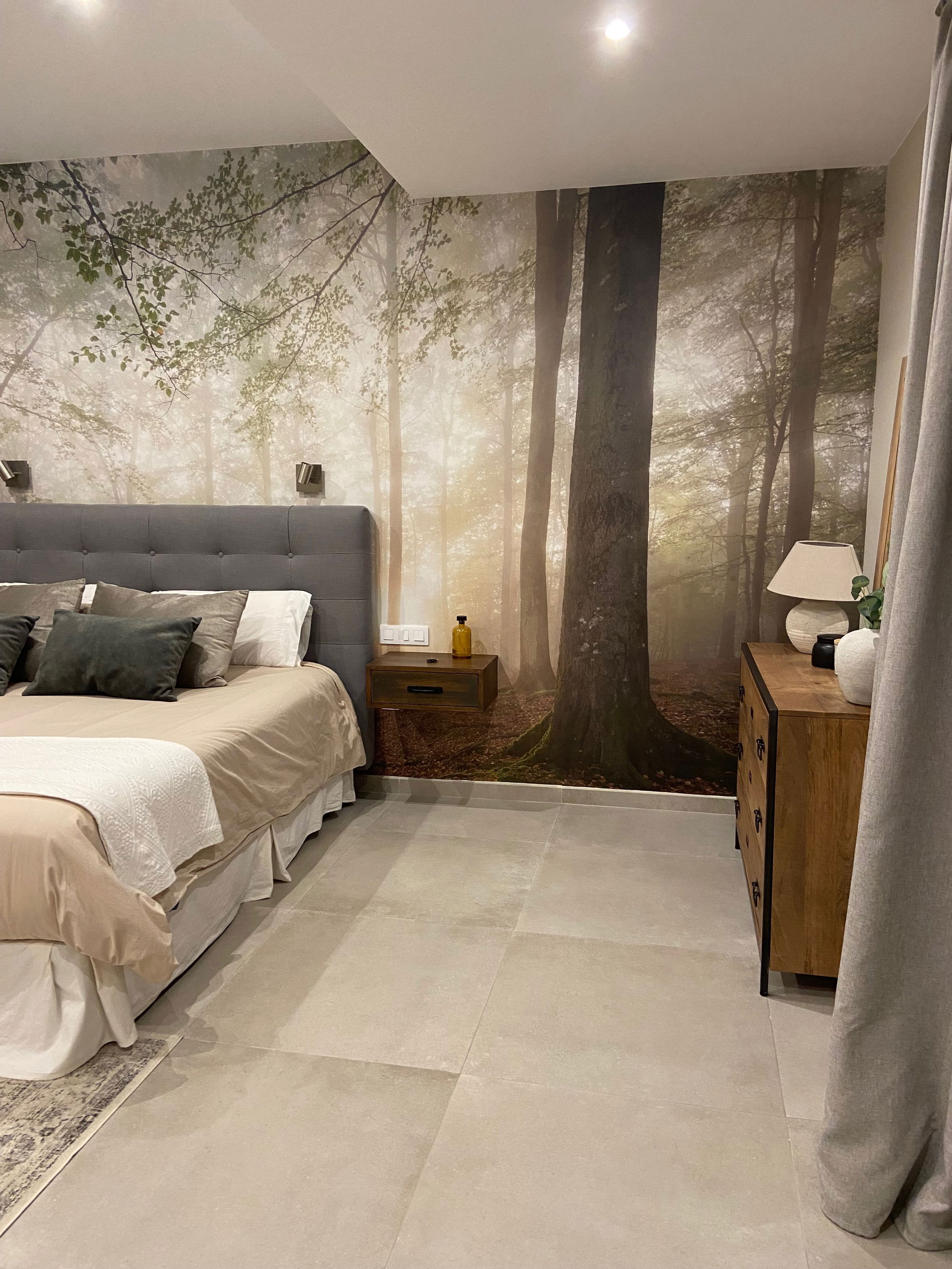 Schlafzimmer mit Wald-Wandbild, grauem getuftetem Kopfteil und Holzkommode.
