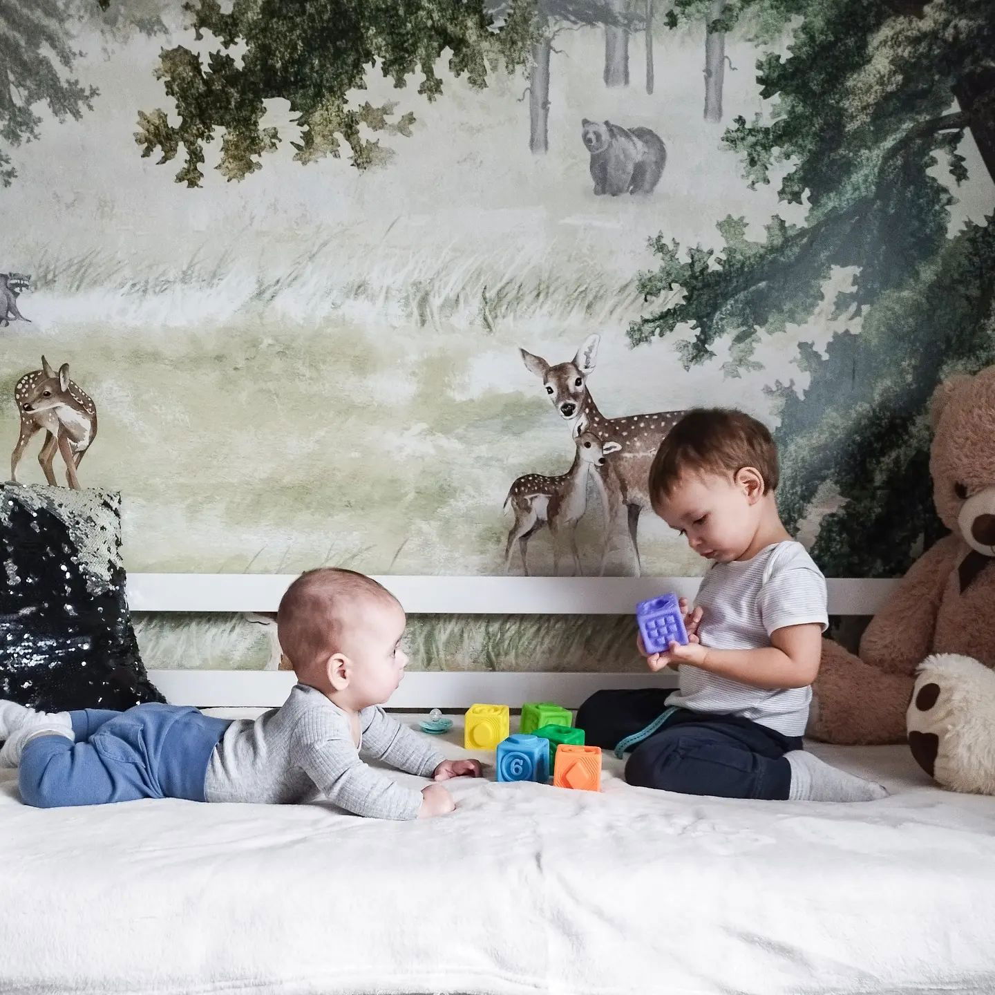 Deux jeunes enfants jouent avec des blocs colorés sur une couverture blanche devant un mur sur le thème de la forêt.