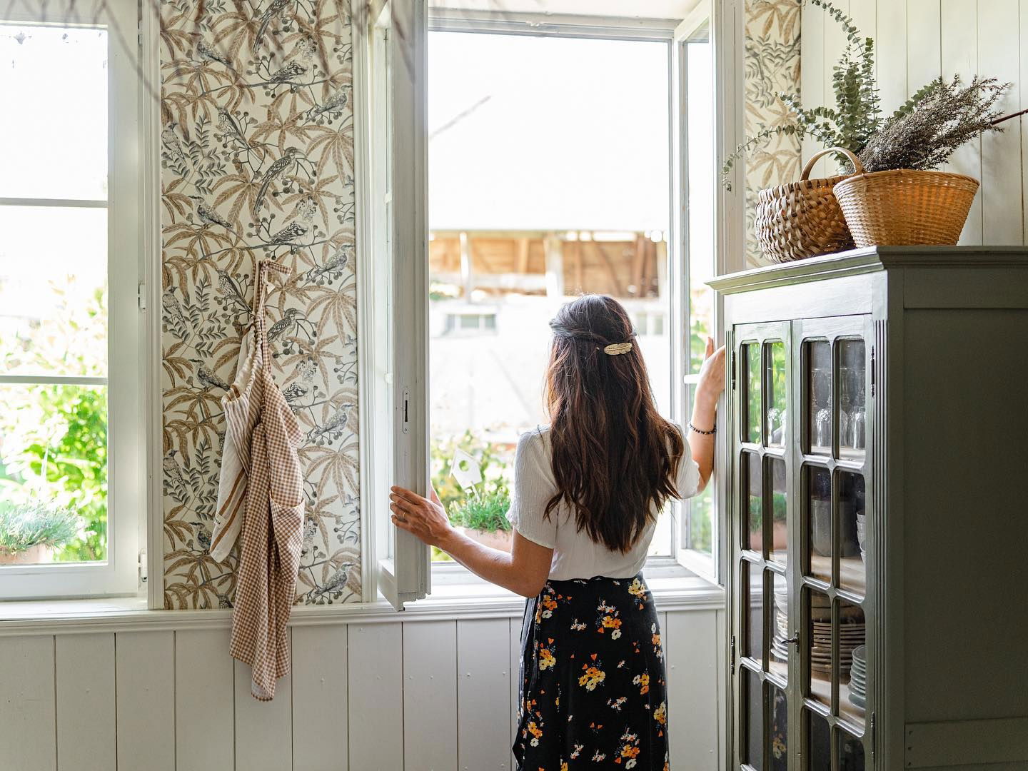 Una mujer abre una ventana en una cocina de estilo vintage con papel tapiz floral.