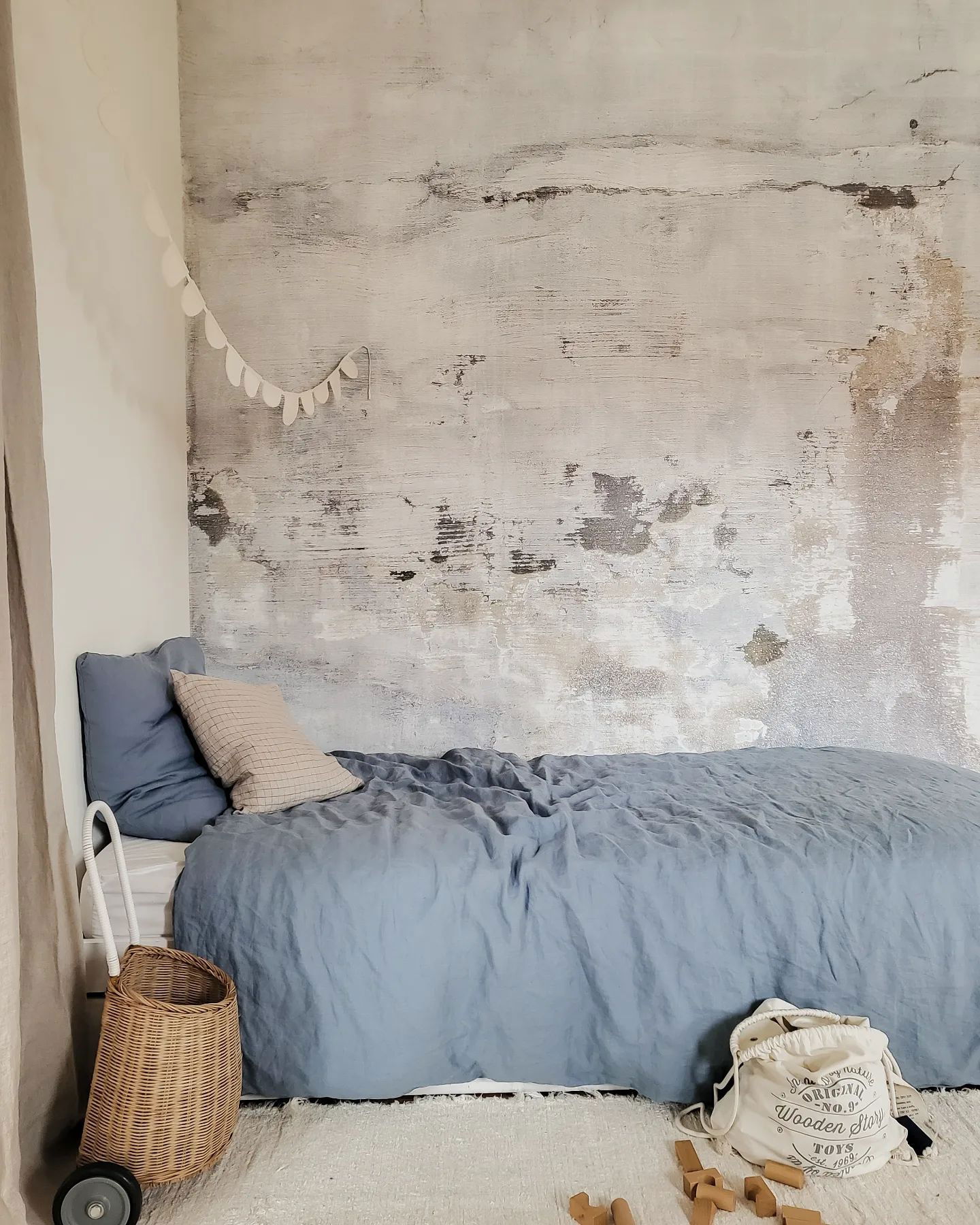 Kinderkamer met blauw linnen beddengoed, getextureerde muur en "Wooden Story" speelgoedzak.