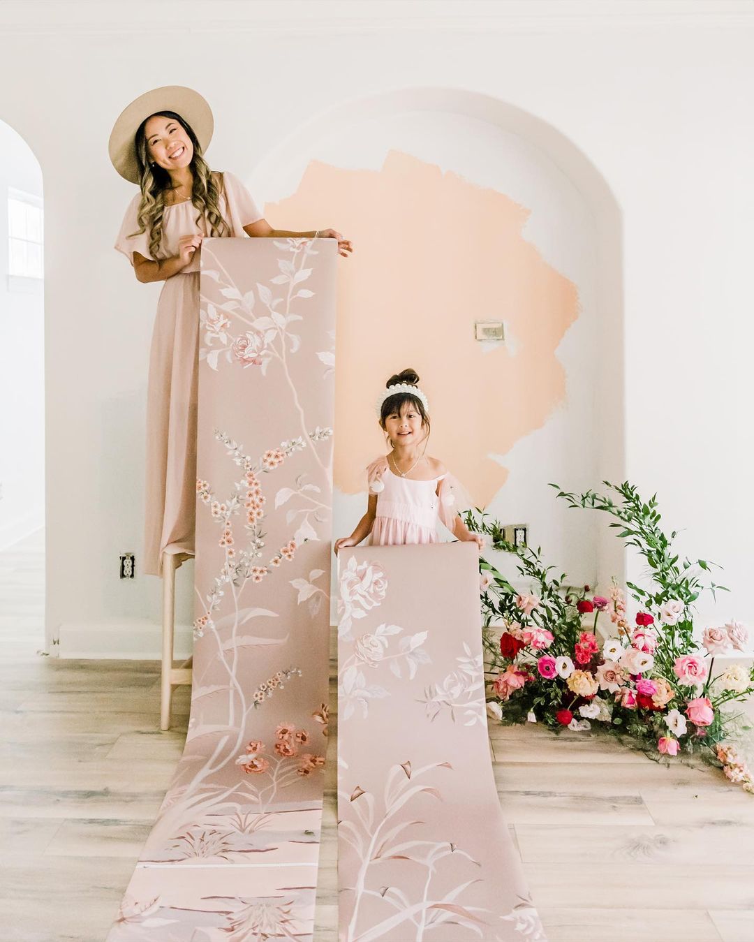 Woman and child holding floral wallpaper rolls in front of a painted archway with floral arrangement.