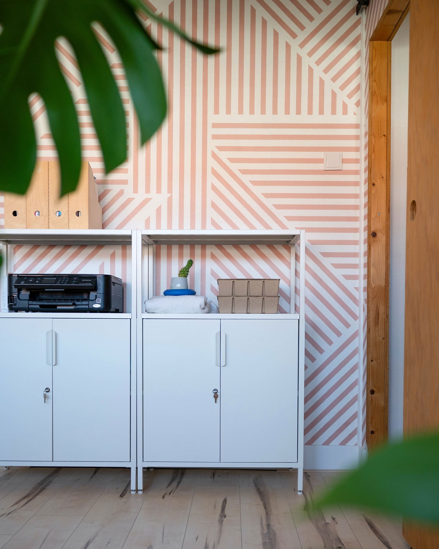 Office interior with white metal cabinets, shelves, and pink striped geometric wallpaper.