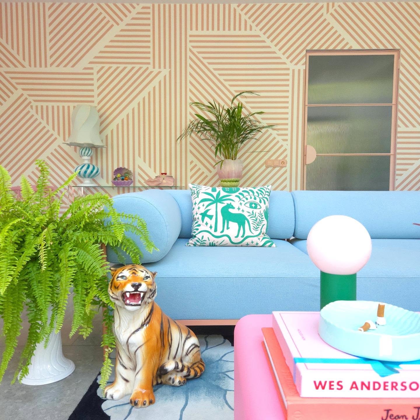 A colorful room with a blue couch, an orange tiger statue, and a fern. Beige striped wallpaper, pink coffee table.