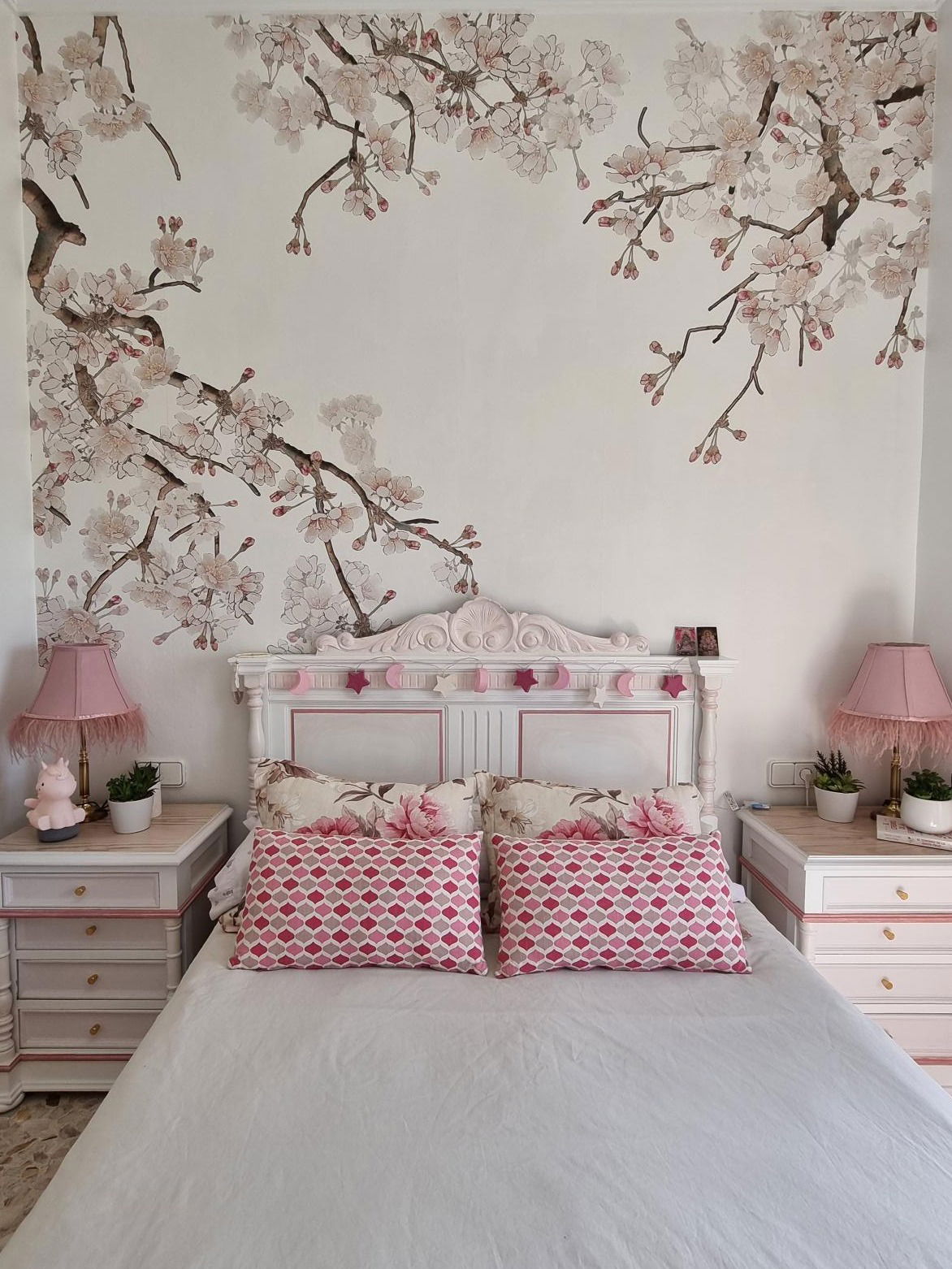 Bedroom with white and pink bed, floral pillows, and cherry blossom wallpaper.