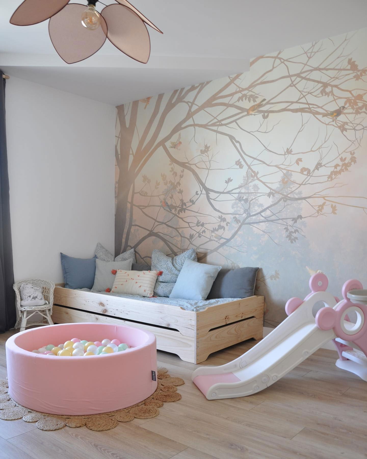 Playroom with a light wood daybed, pink ball pit, and white and pink slide. Tree mural on the wall.