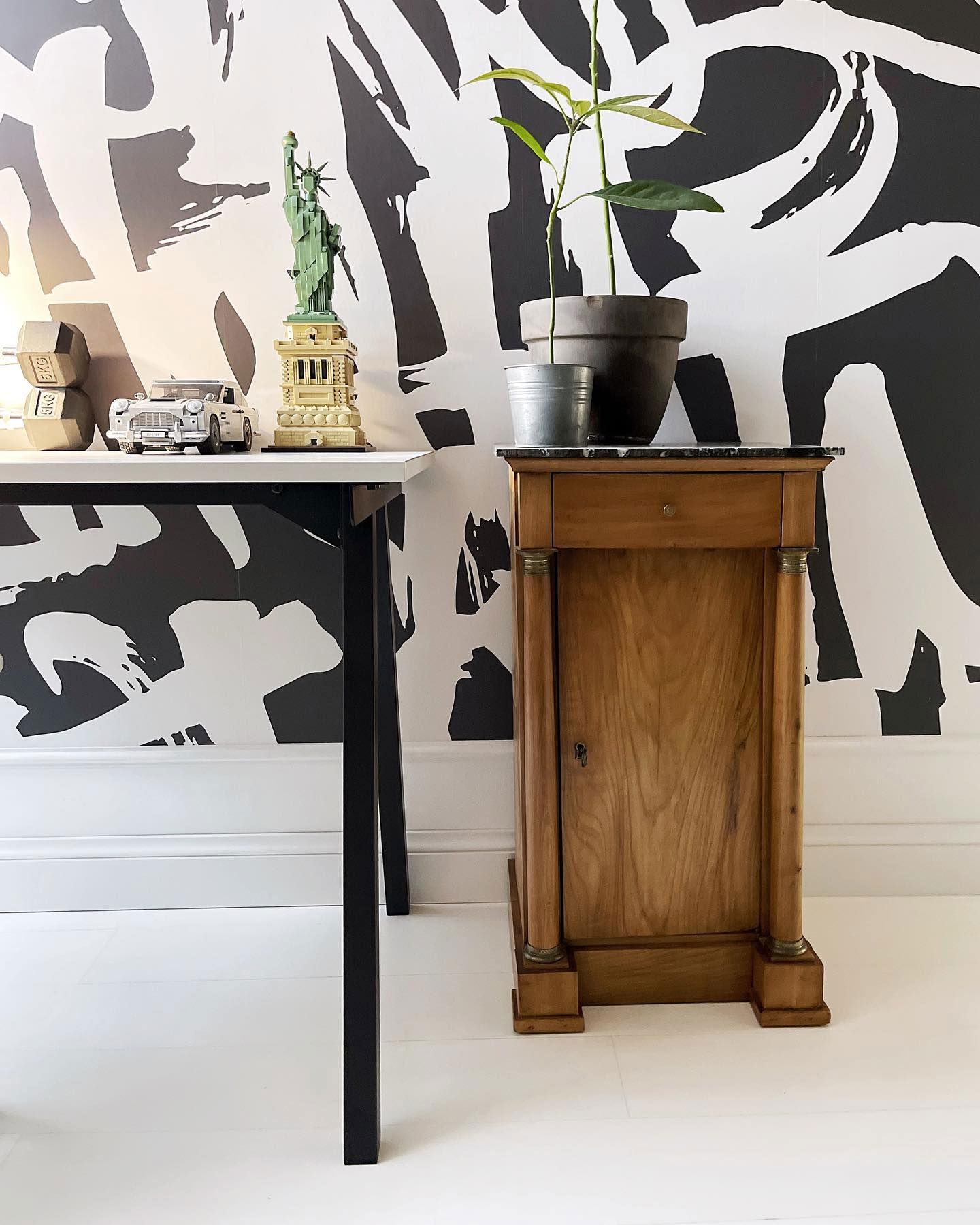 Interno con arte murale in bianco e nero, scrivania con statua della Libertà Lego e mobile in legno con pianta in vaso.