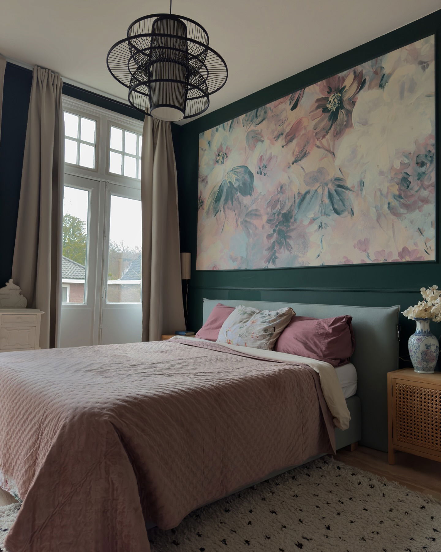 Slaapkamer met roze quilt, bloemenkunst aan de muur en zwarte hanglamp.