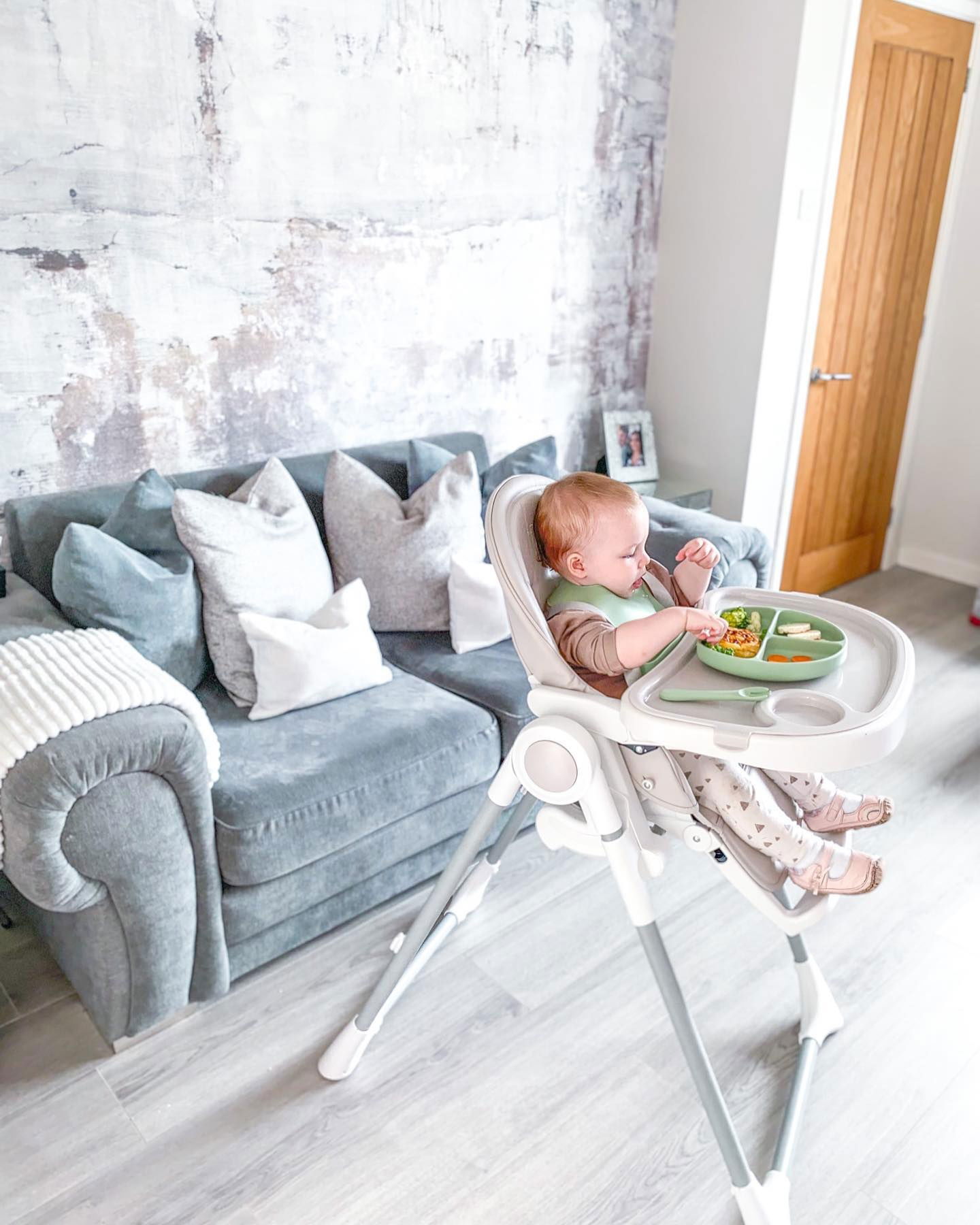 Baby in kinderstoel eet voedsel van een verdeeld groen bord met broccoli, wortels en courgette.