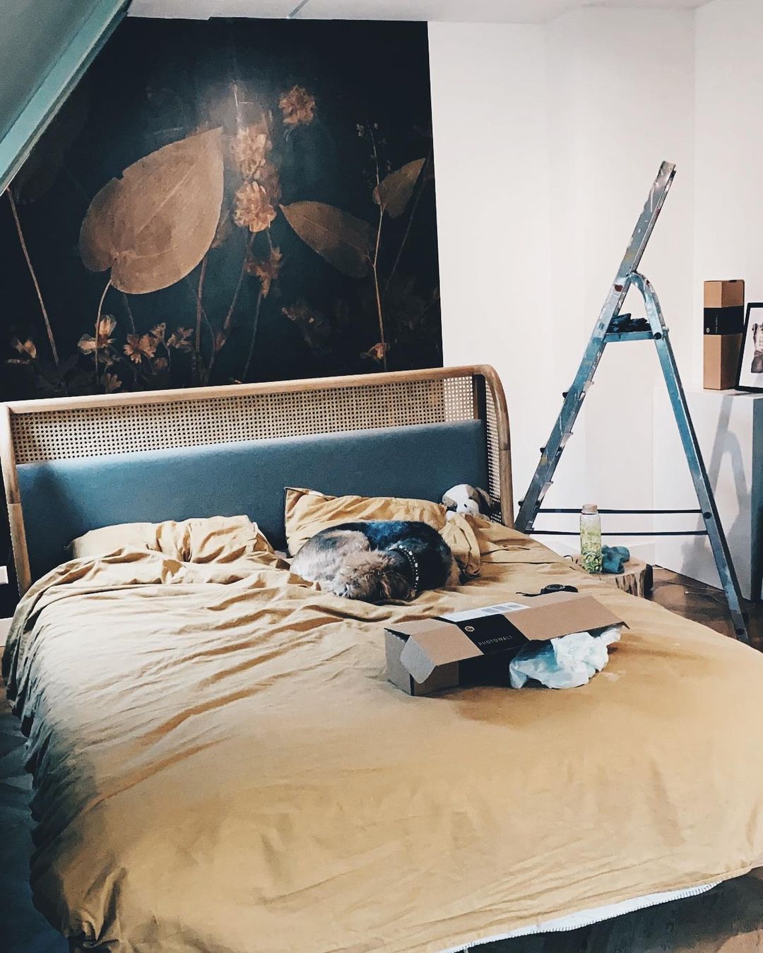 Dormitorio con ropa de cama color mostaza, perro durmiendo y caja de Photowall. Escalera y arte floral en la pared al fondo.