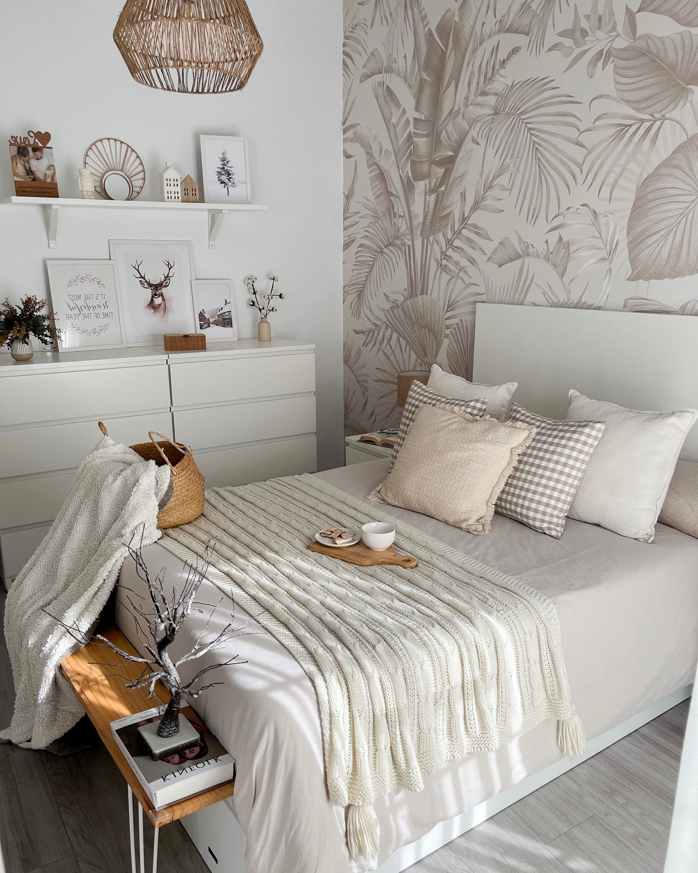 Bedroom with white bed, neutral pillows, and chunky knit throw. Tropical wallpaper and woven pendant light add texture.