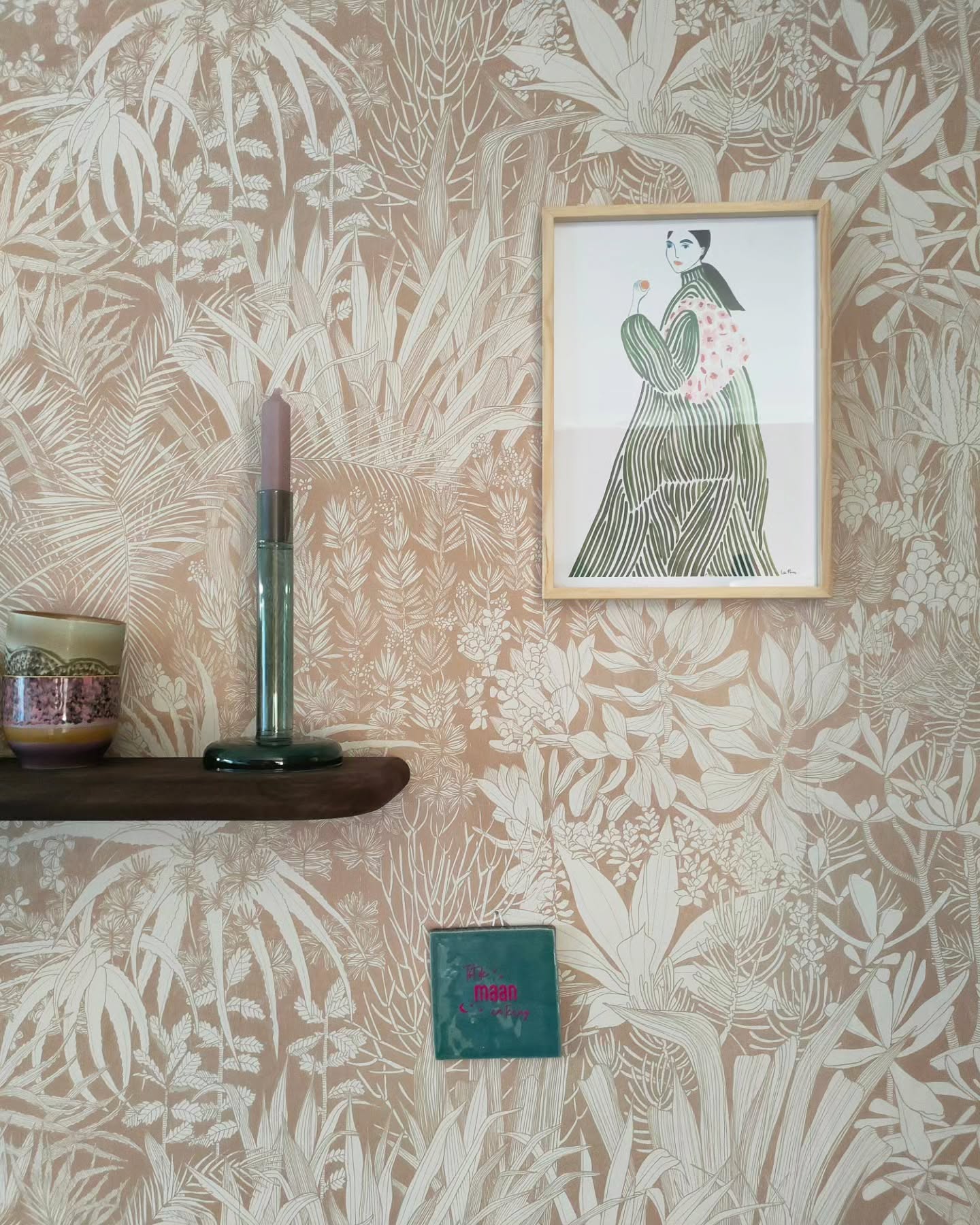 Pared interior con papel tapiz floral, estante con vela, taza, libro y arte enmarcado.