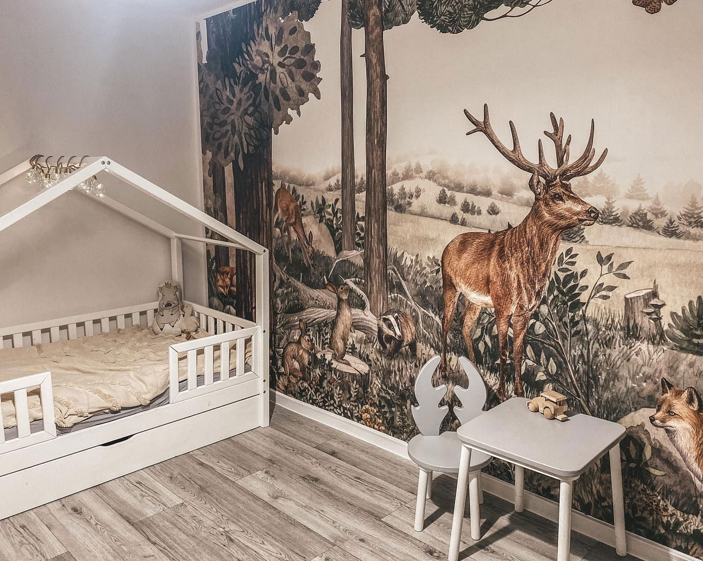 Child's room with woodland mural, white house-frame bed, and gray table with antler chair.