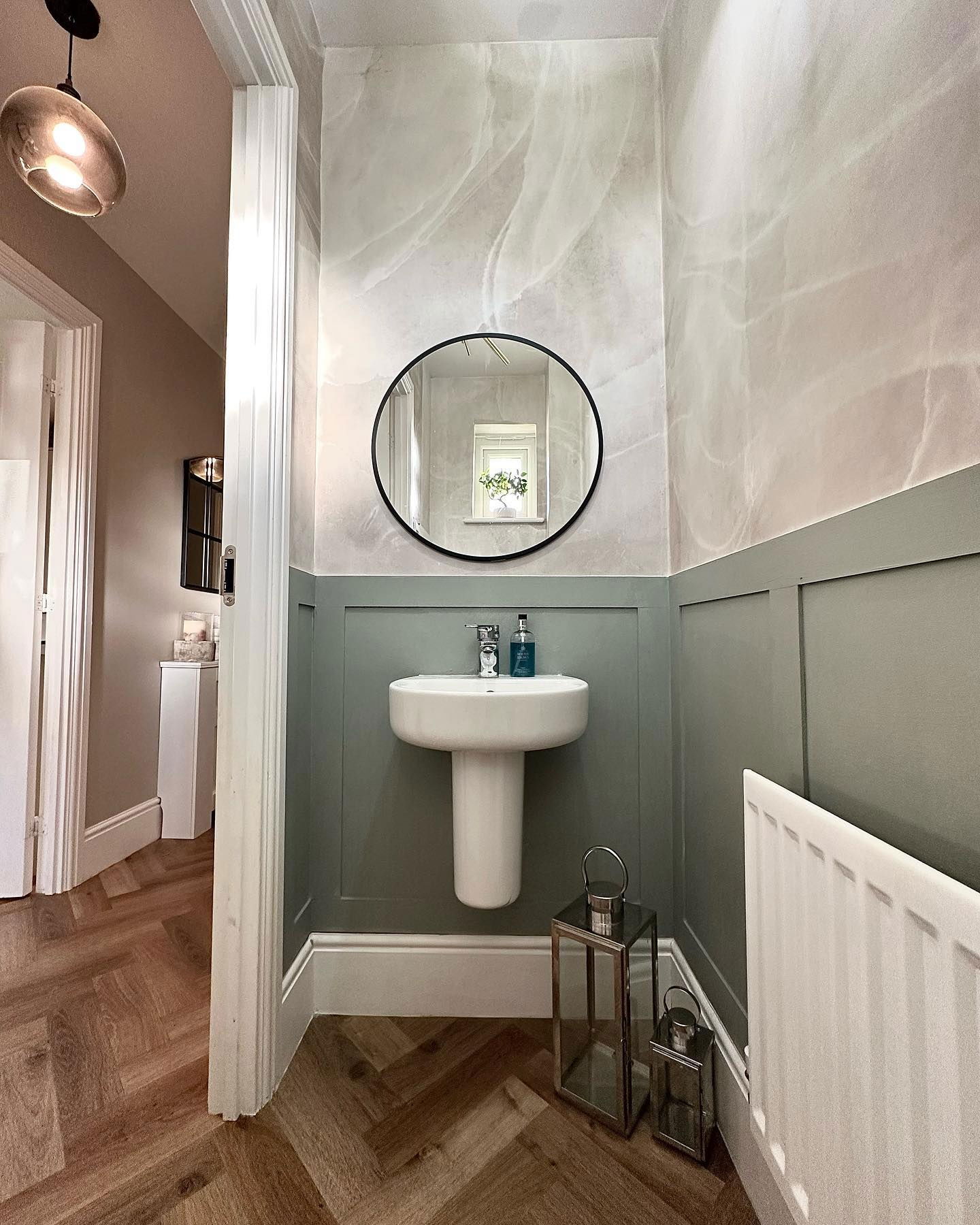 Bagno con lavabo a colonna bianco, specchio rotondo e pareti rivestite di pannelli verdi. Pavimenti in legno a spina di pesce.
