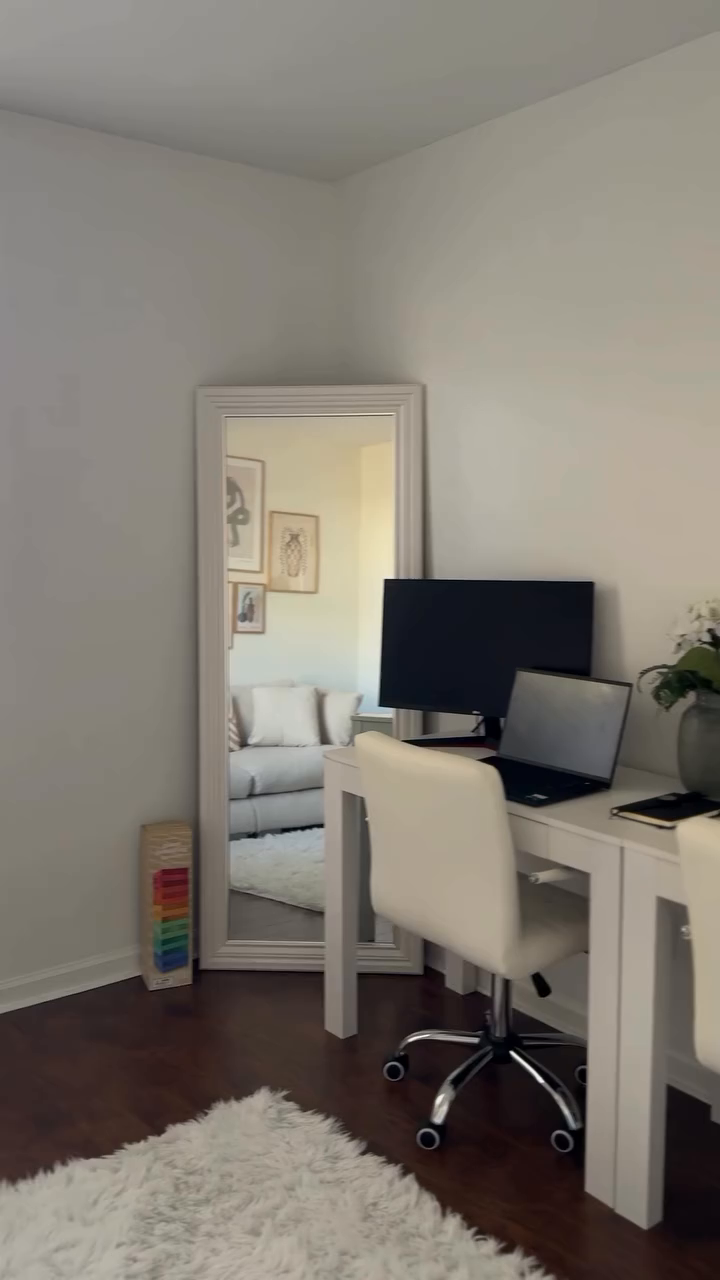 Bright home office with white desk, chair, computer, and full-length mirror reflecting a living room.