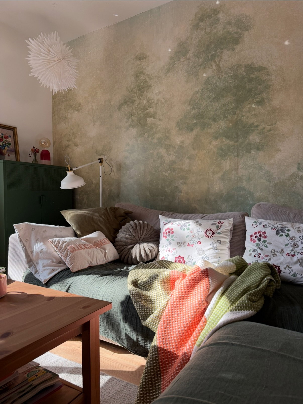 Soffa med blommiga kuddar, strukturerade plädar och en konstnärlig grön muralmålning på väggen.