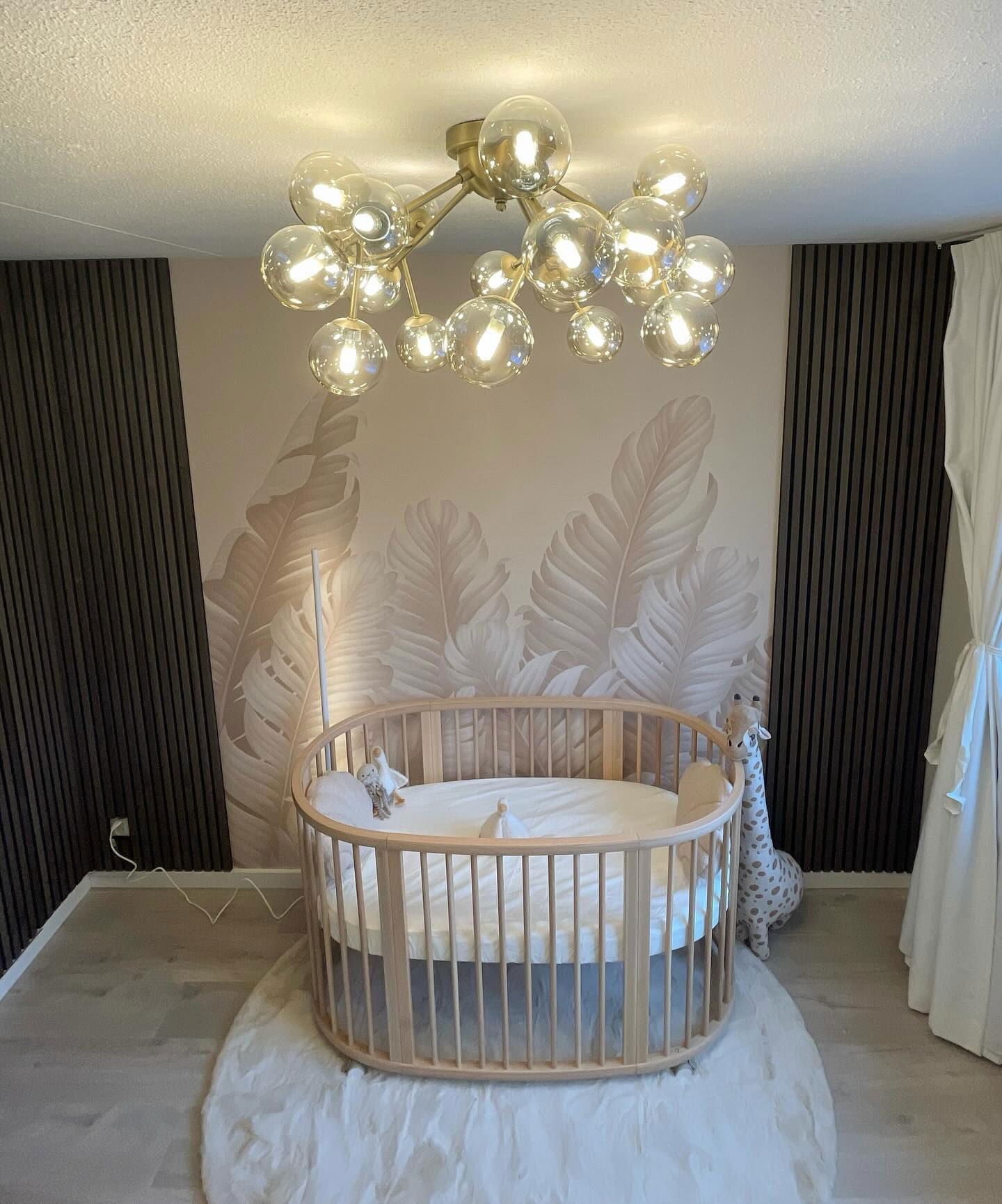 Kinderkamer met rond houten wiegje op wit vloerkleed, behang met bladmotief en bubbellamp.