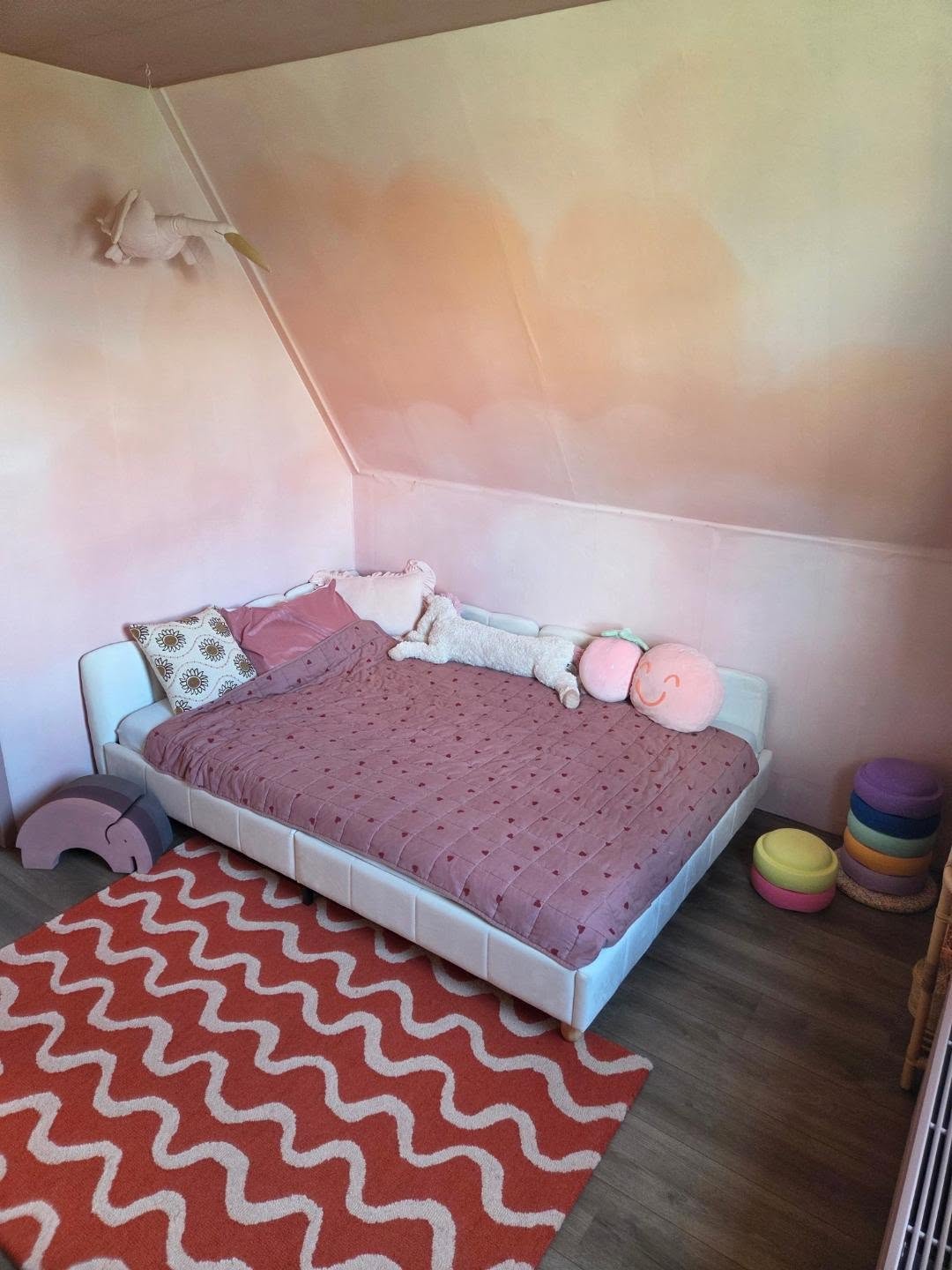 White bed with pink heart-patterned quilt, plush toys, and a wavy orange rug in a child's room.
