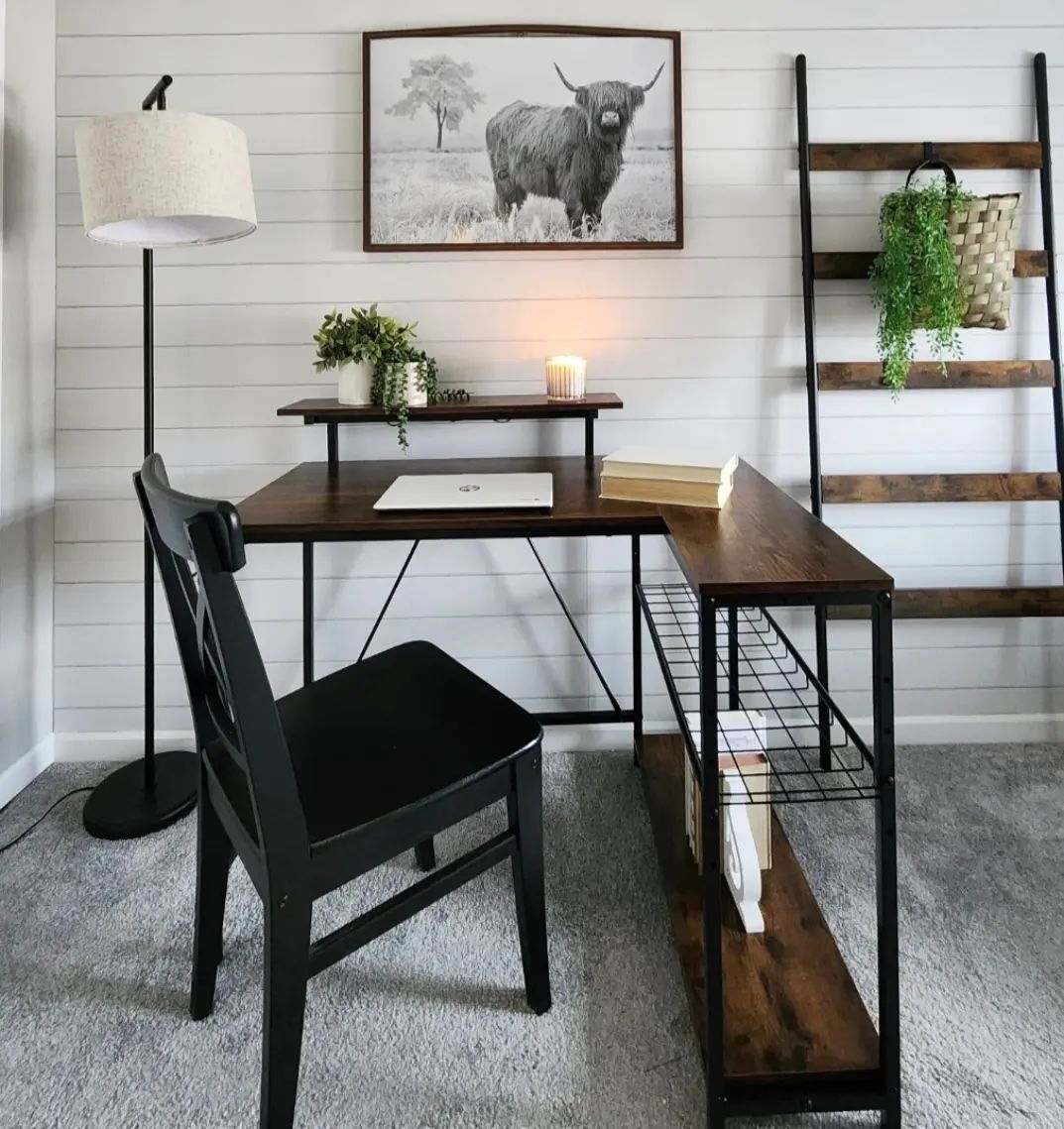Home office setup with wood desk, black chair, and ladder shelf against a white shiplap wall. Highland cow art.