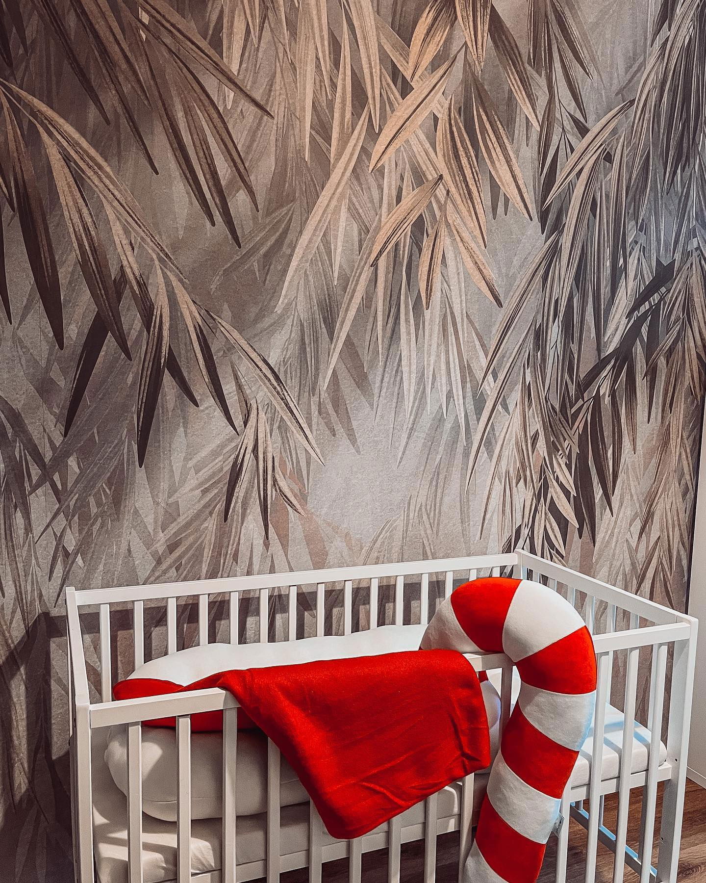 White baby crib with red blanket and candy cane pillow against a leaf-patterned wall.