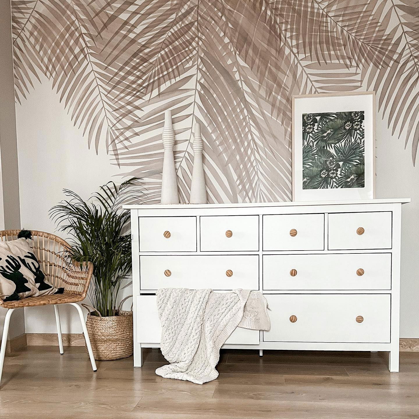 Interior de dormitorio con tocador blanco, estampado tropical, silla de mimbre y papel tapiz de hojas de palma.