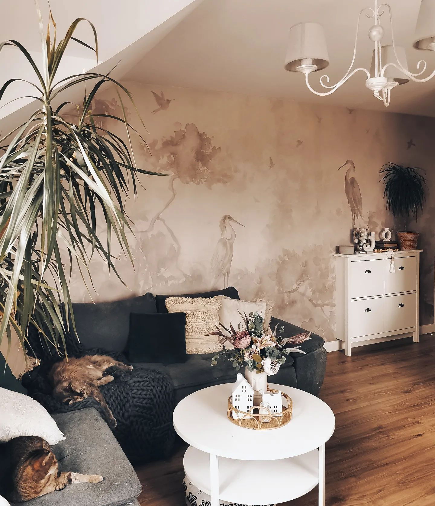 Cozy living room with gray sofa, cats, and heron-themed wallpaper. A white table holds a floral arrangement.