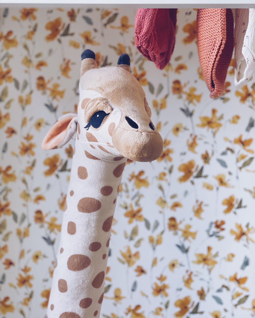 Stuffed giraffe toy with brown spots, against floral wallpaper and hanging baby clothes.