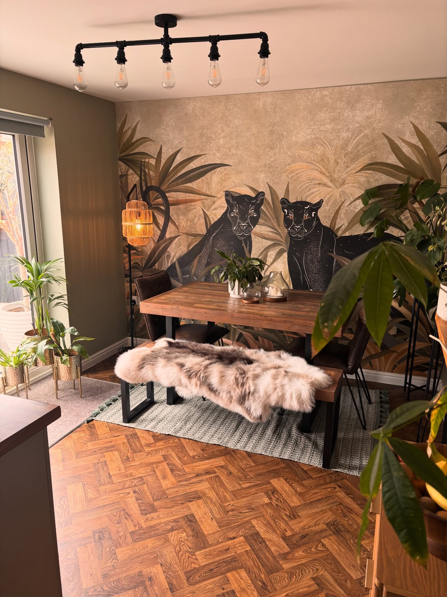 Dining room with jungle wallpaper of two black panthers. Wooden table, bench with light faux fur throw, black pipe chandelier, plants, herringbone floor.
