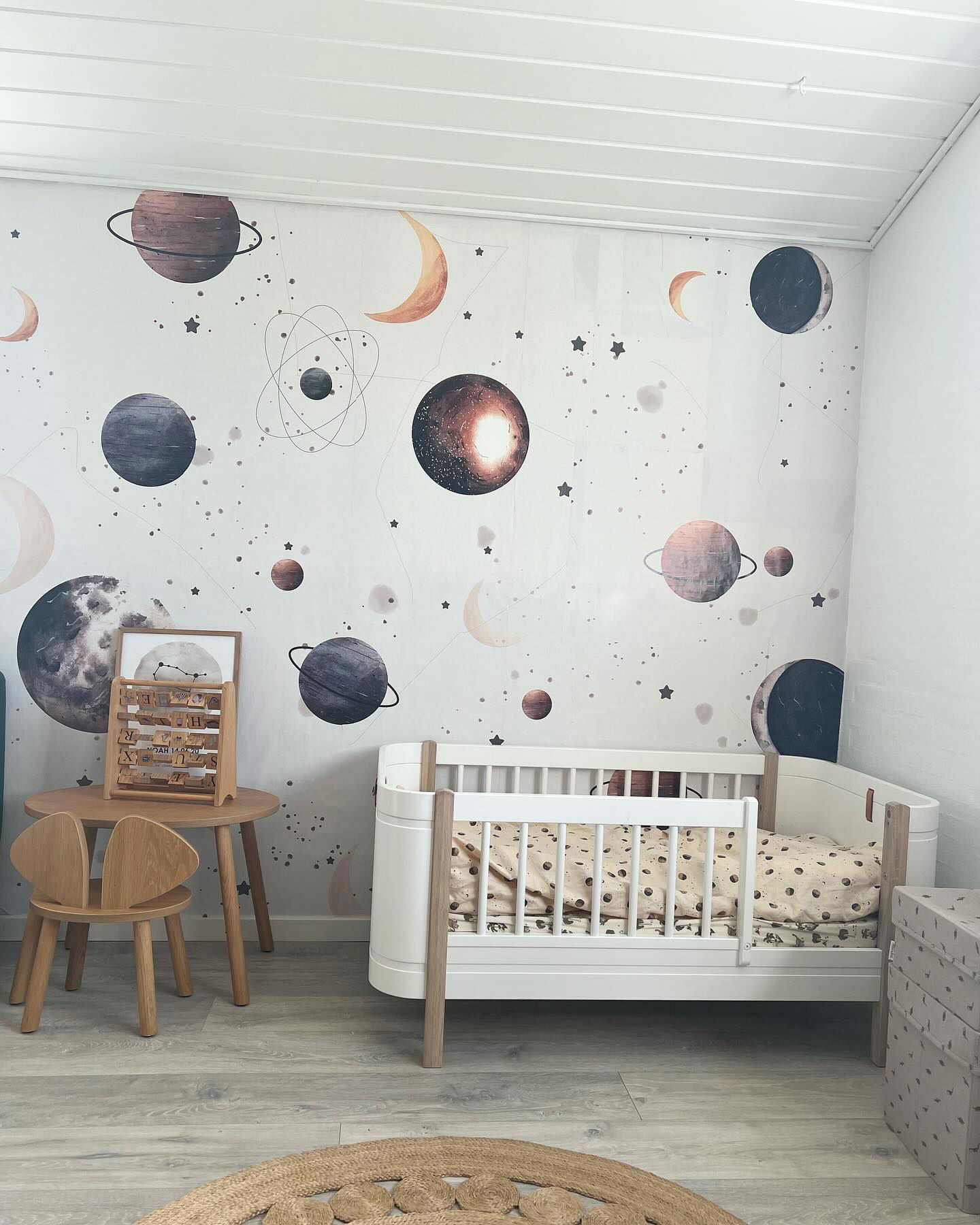 Kinderzimmer mit weißer Wiege, Holzstuhl und Tisch sowie Weltraumtapete.