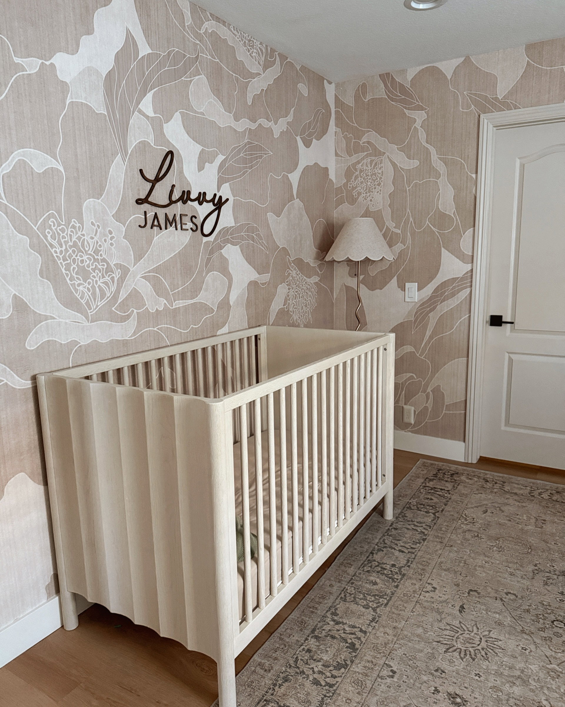 White wooden crib in a nursery. Beige and white floral wallpaper, a dark wooden 'Livvy James' sign above the crib. Patterned rug.