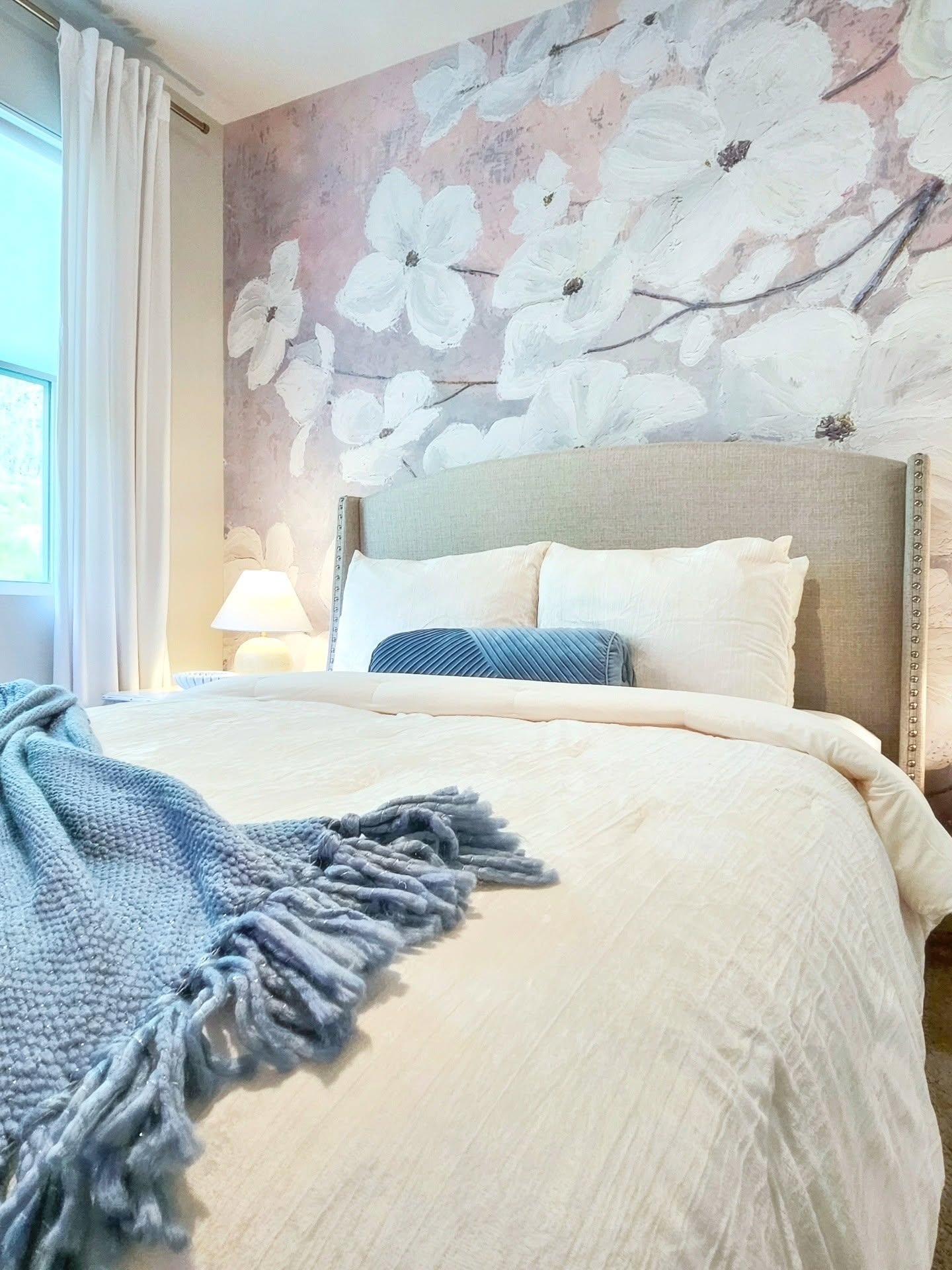 Bedroom with a bed, light beige bedding, a blue knit throw, and a beige upholstered headboard. A pink accent wall with large white floral art is behind the bed.