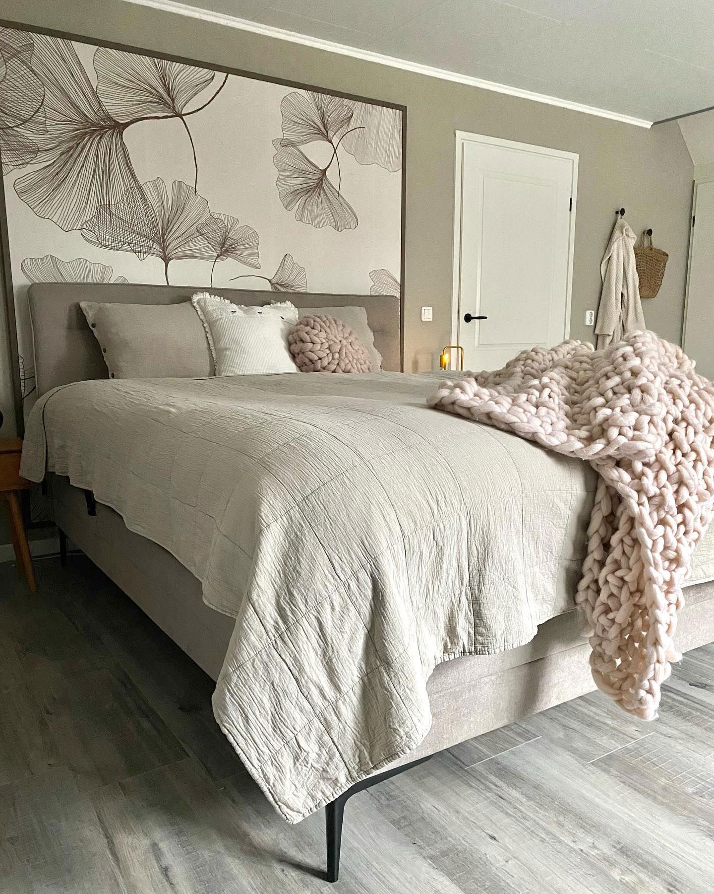 Chambre avec lit beige, courtepointe en lin et couverture en grosse maille. Art mural floral au-dessus de la tête de lit.
